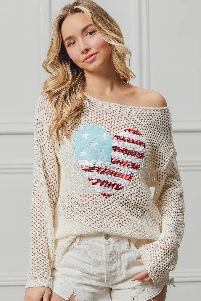 Woman wearing a cream sweater with a heart-shaped American flag design against a white wall.