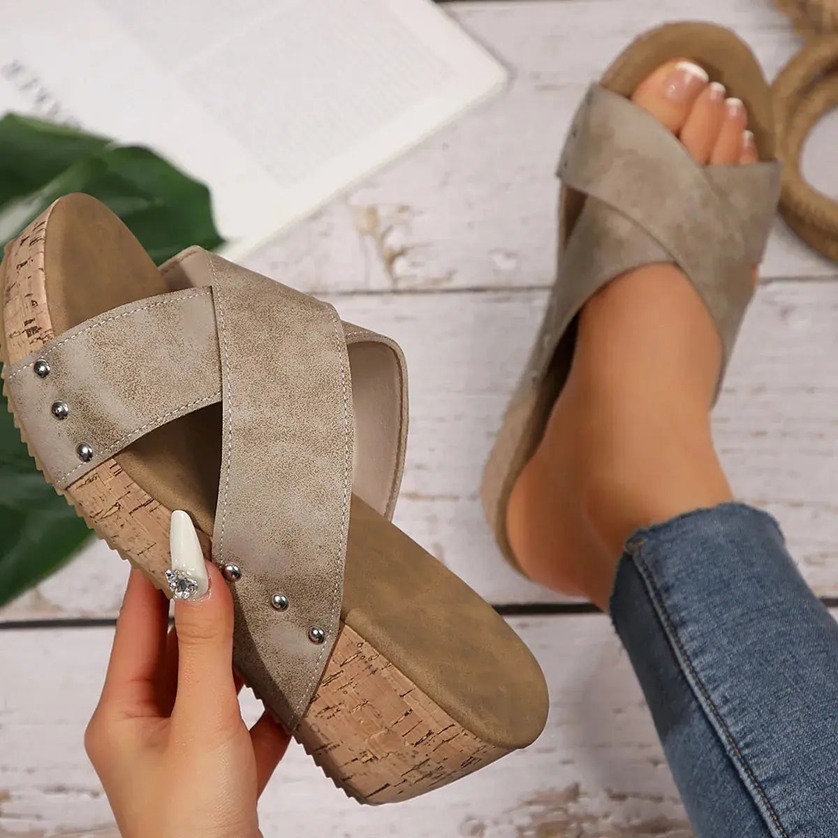 Beige sandals with cork sole held by a hand on a light wooden floor.