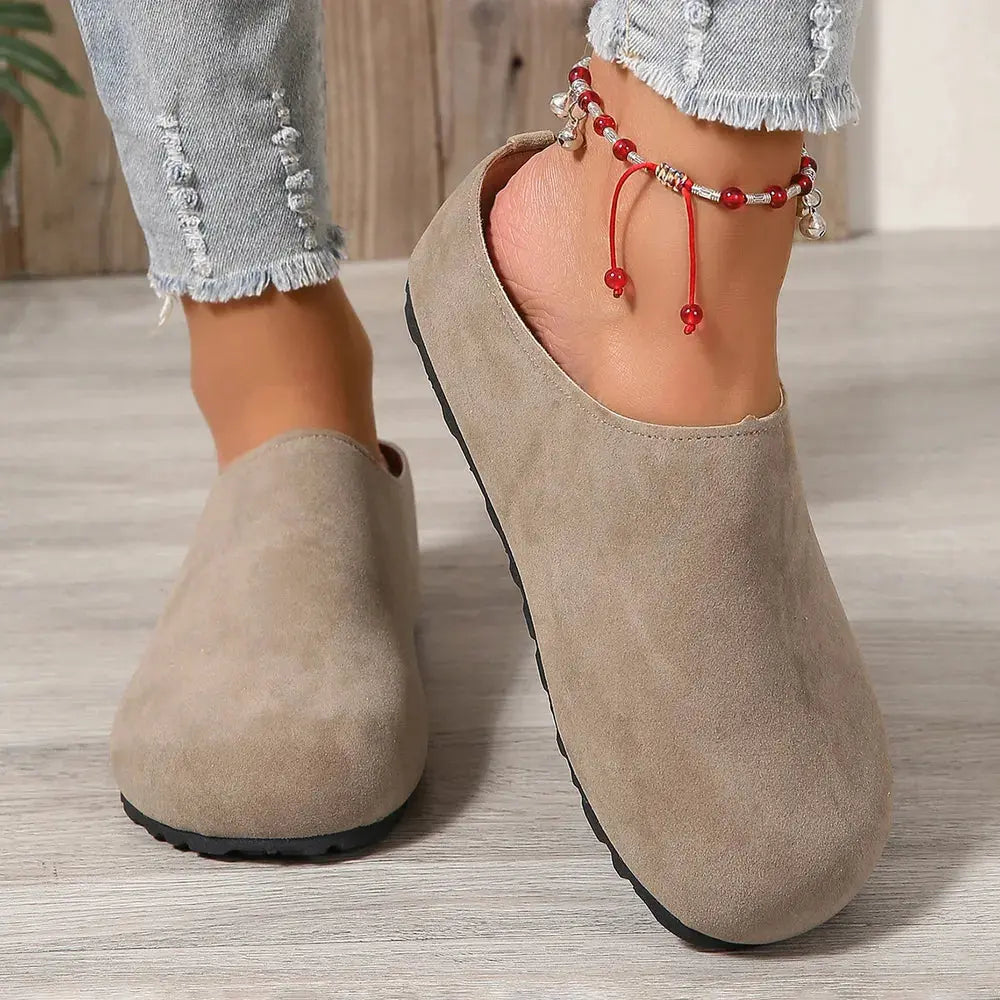 Beige suede clogs worn with light blue jeans on a wooden floor.