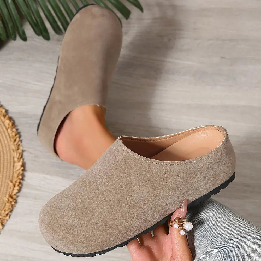 Beige suede clogs worn by a person on a light wooden floor.