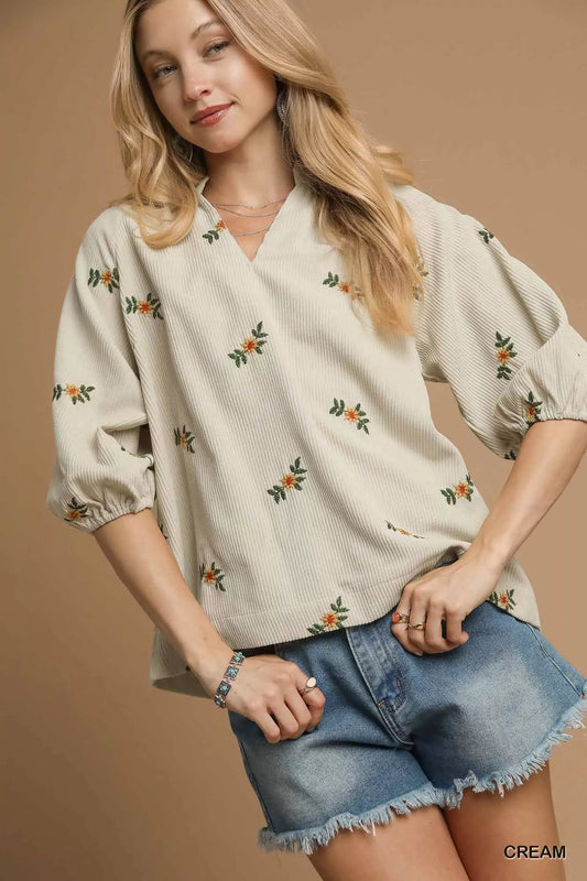 Woman wearing a cream blouse with floral patterns and denim shorts on a beige background