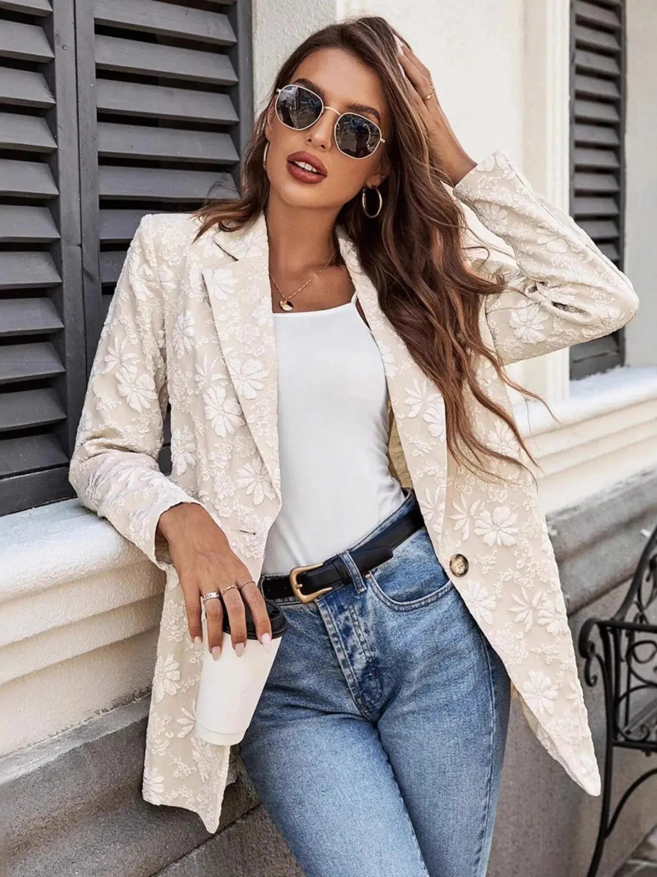 Woman wearing a beige floral coat, white top, and blue jeans outdoors.
