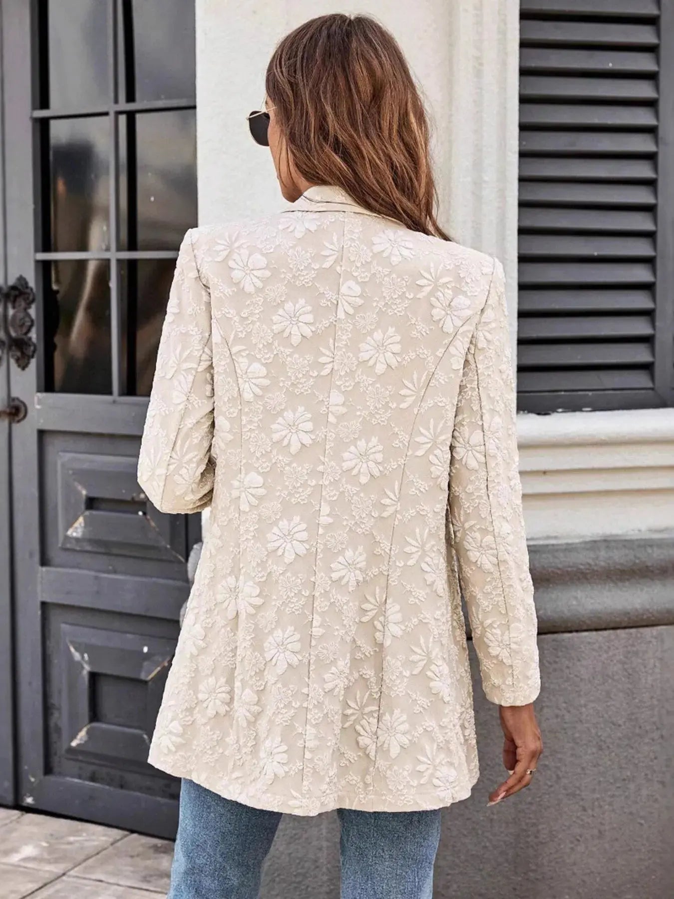 Person wearing a beige floral-patterned blazer standing in front of a building entrance.