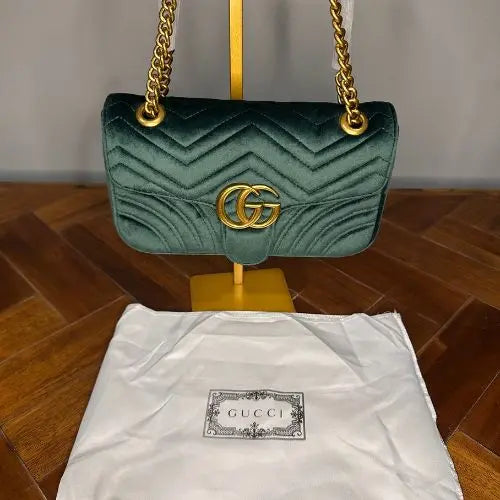 Green velvet handbag with gold chain and GG logo on a wooden surface hanging over a white dustbag.