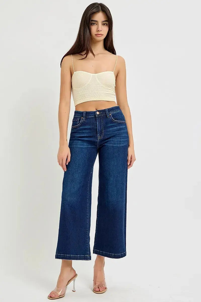 Woman wearing a cream spaghetti strap top and cropped, wide leg blue jeans on a white background
