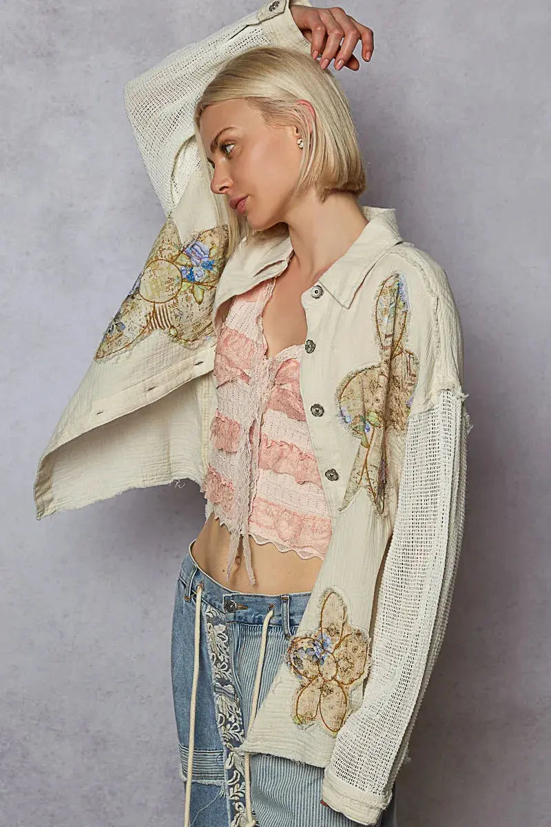 Woman wearing a cream-colored stylish button down that has floral patches and mesh sleeves.