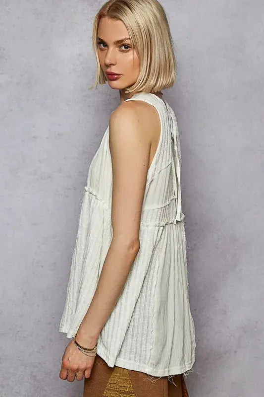 Woman standing to show the side view of a white flowy sleeveless top against a gray background.