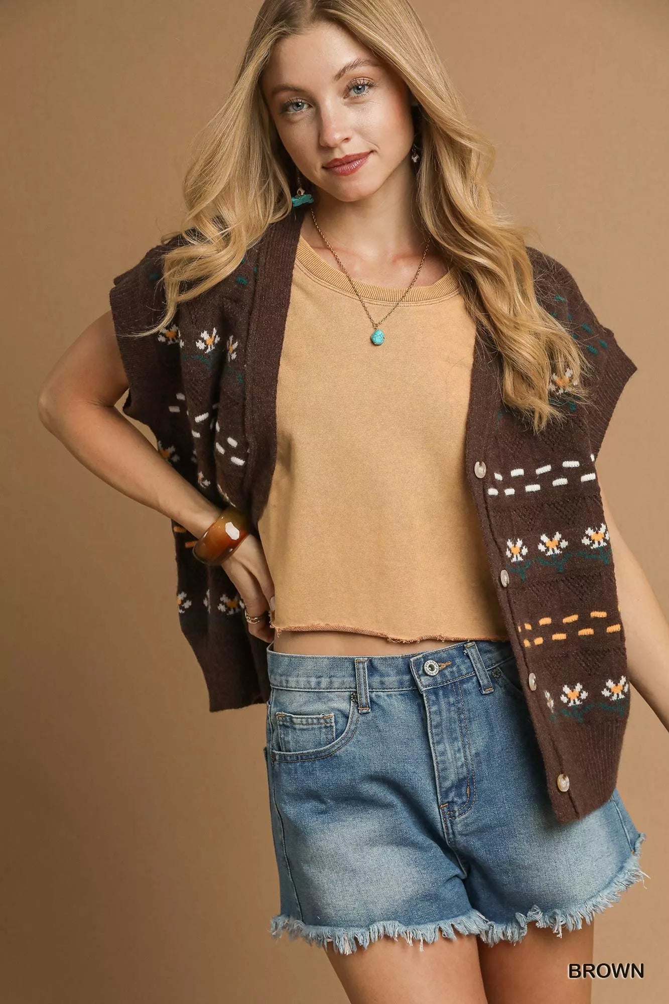 Woman wearing a patterned vest over a beige top and denim shorts on a brown background
