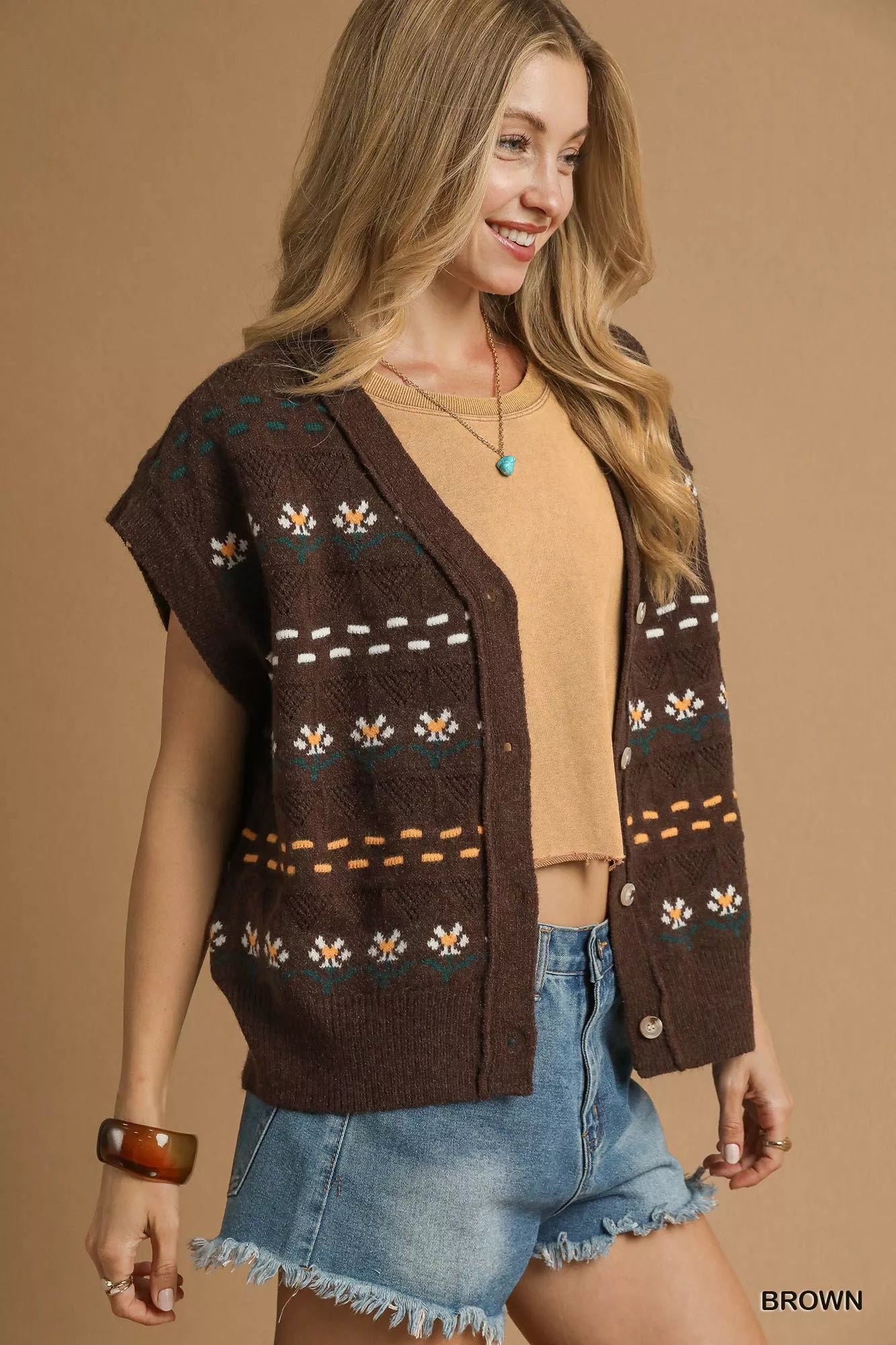 Woman wearing a patterned vest over a beige top and denim shorts on a brown background
