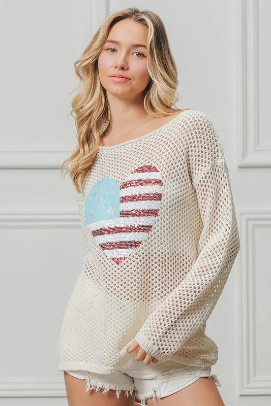 Woman wearing a beige sweater with an American flag heart design against a light gray wall.