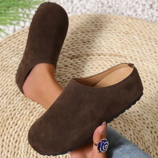 Brown suede clogs worn by a person sitting on a woven chair.