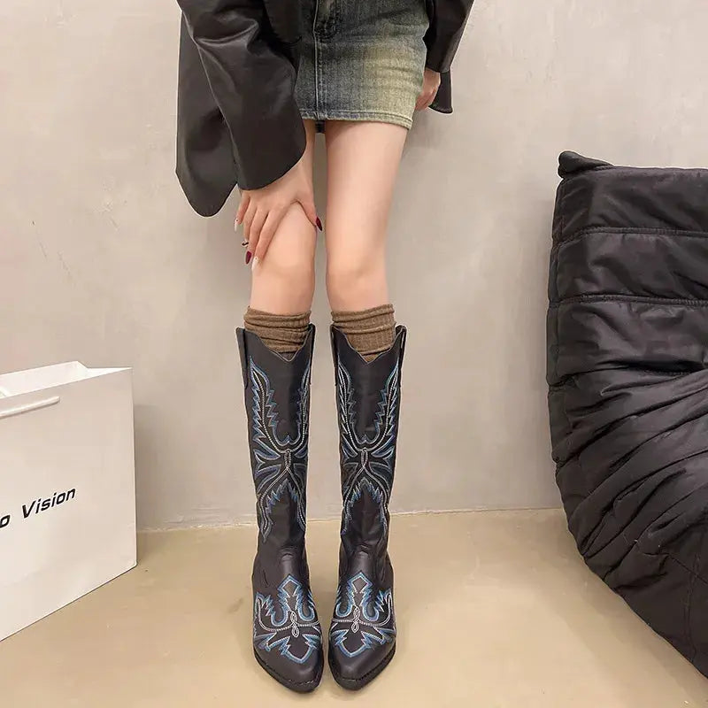 Person wearing black cowboy boots with blue designs, standing against a neutral background.