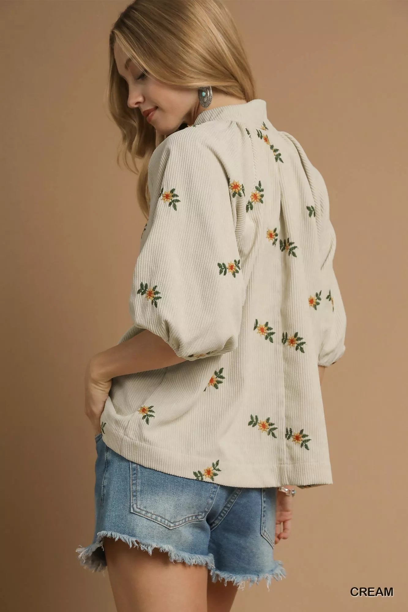 Woman wearing a cream-colored blouse with floral patterns and denim shorts on a beige background
