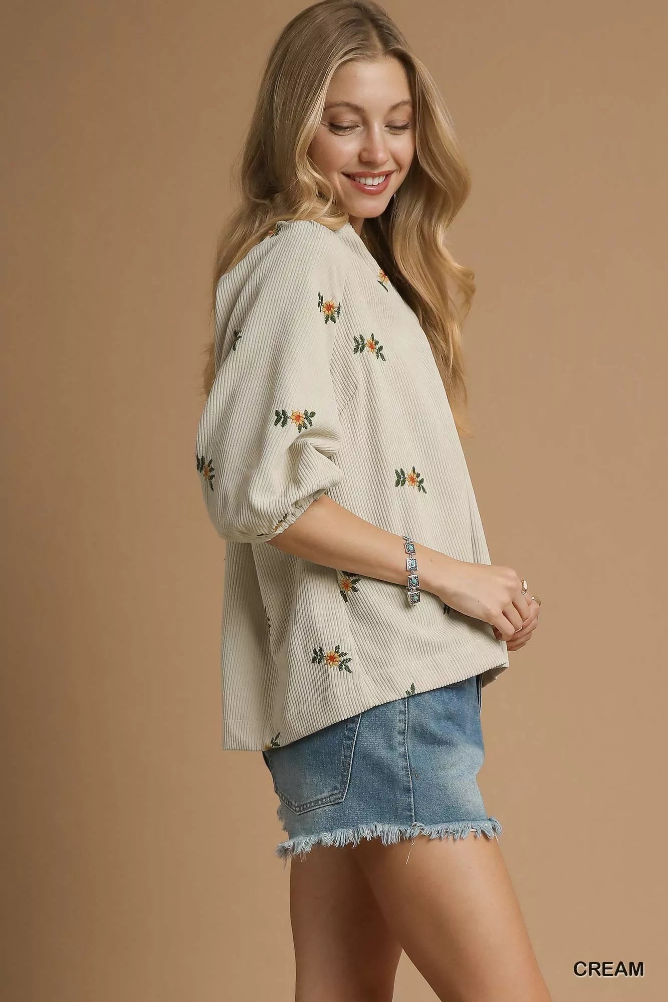 Woman wearing a cream-colored embroidered top and denim shorts on a beige background