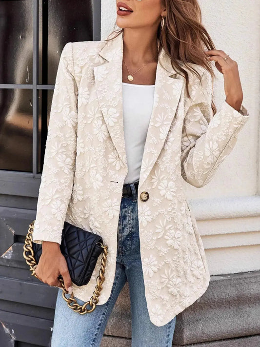 Woman wearing a beige floral blazer, white top, and blue jeans, holding a black handbag with gold chain.