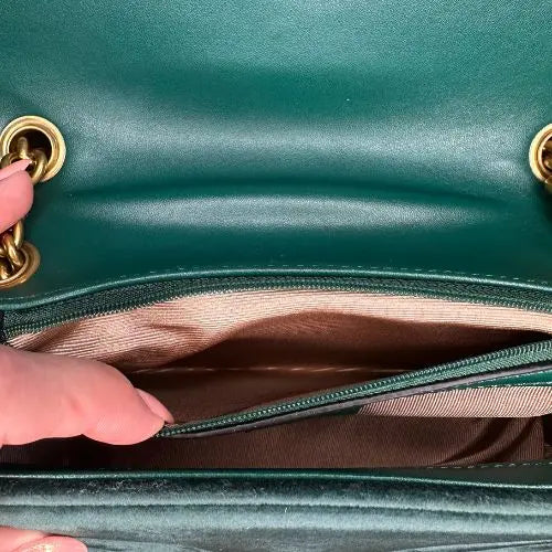 Close-up of a green handbag with gold hardware, showing a hand opening its zippered compartment.