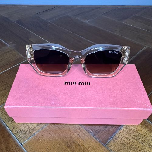 Sunglasses with clear frame and dark lenses on a pink Miu Miu box.