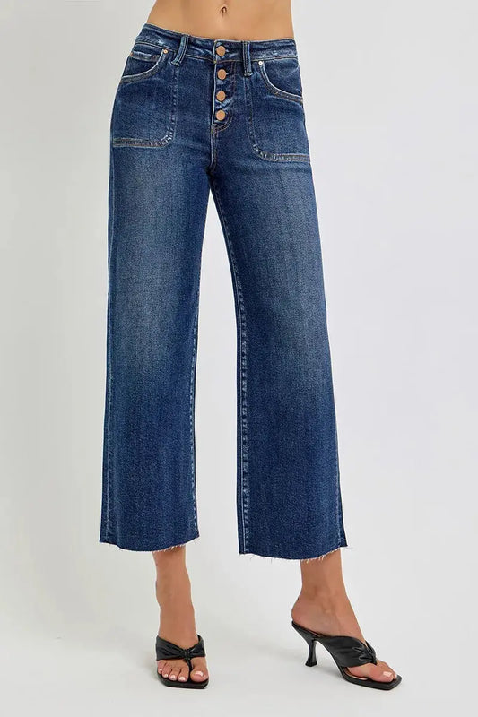 Cropped blue jeans with button closure worn by a person with black heeled sandals on a gray background.