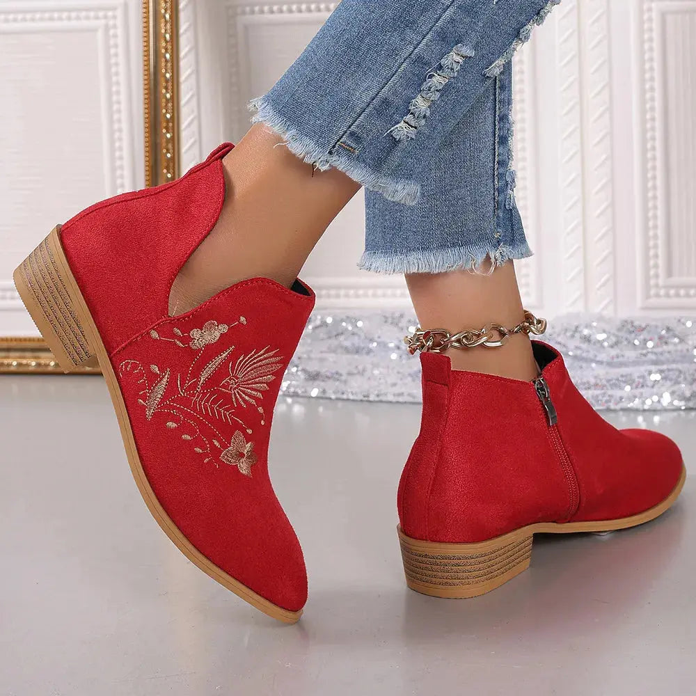 Red ankle boots with floral embroidery worn with blue jeans against a decorative wall.