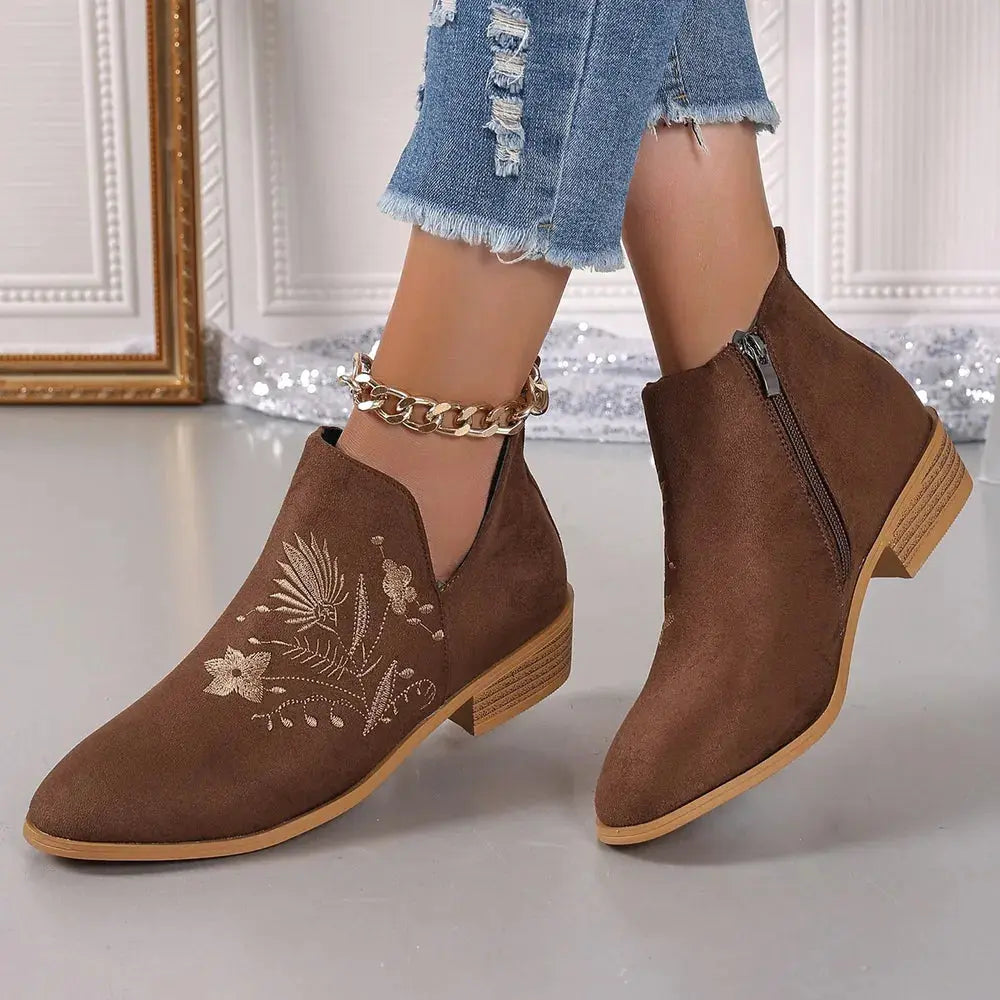 Brown ankle boots with floral embroidery worn by a person on a light gray floor.