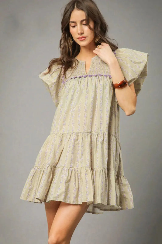 Woman wearing a light green, tiered dress with floral print wand ruffled sleeves against a gray background.