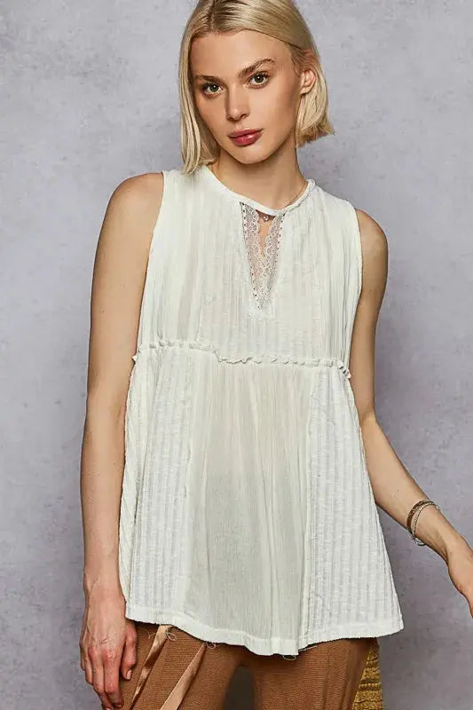 Woman wearing a white sleeveless top with lace details against a gray background.