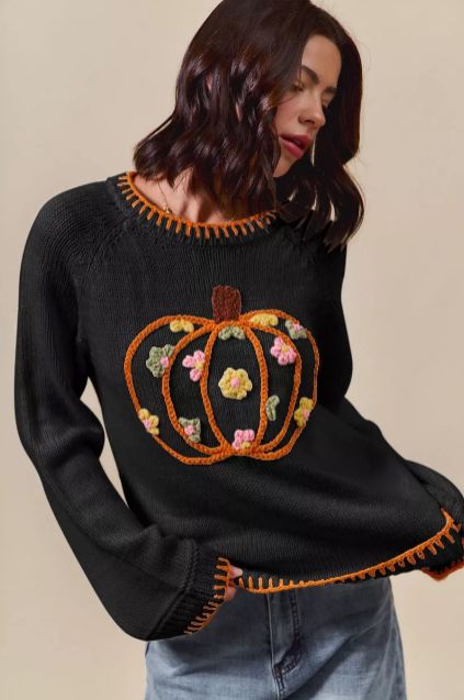 Black knit sweater featuring an embroidered pumpkin with floral details, worn with light wash jeans for a cozy seasonal look. Soft stitching, relaxed drape.